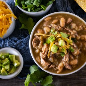 Close top down view of a bowl of pork stew with beans garnished with avocado, onion and cheese.