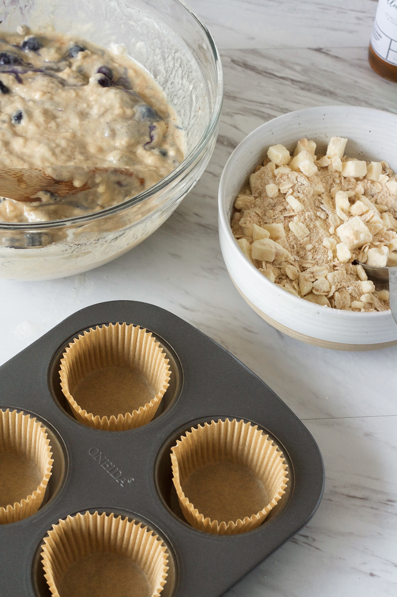 A bowl of blueberry sourdough batter, a bowl of mixed streusel, and a muffin liner filled muffin tin on a counter - Hostess At Heart