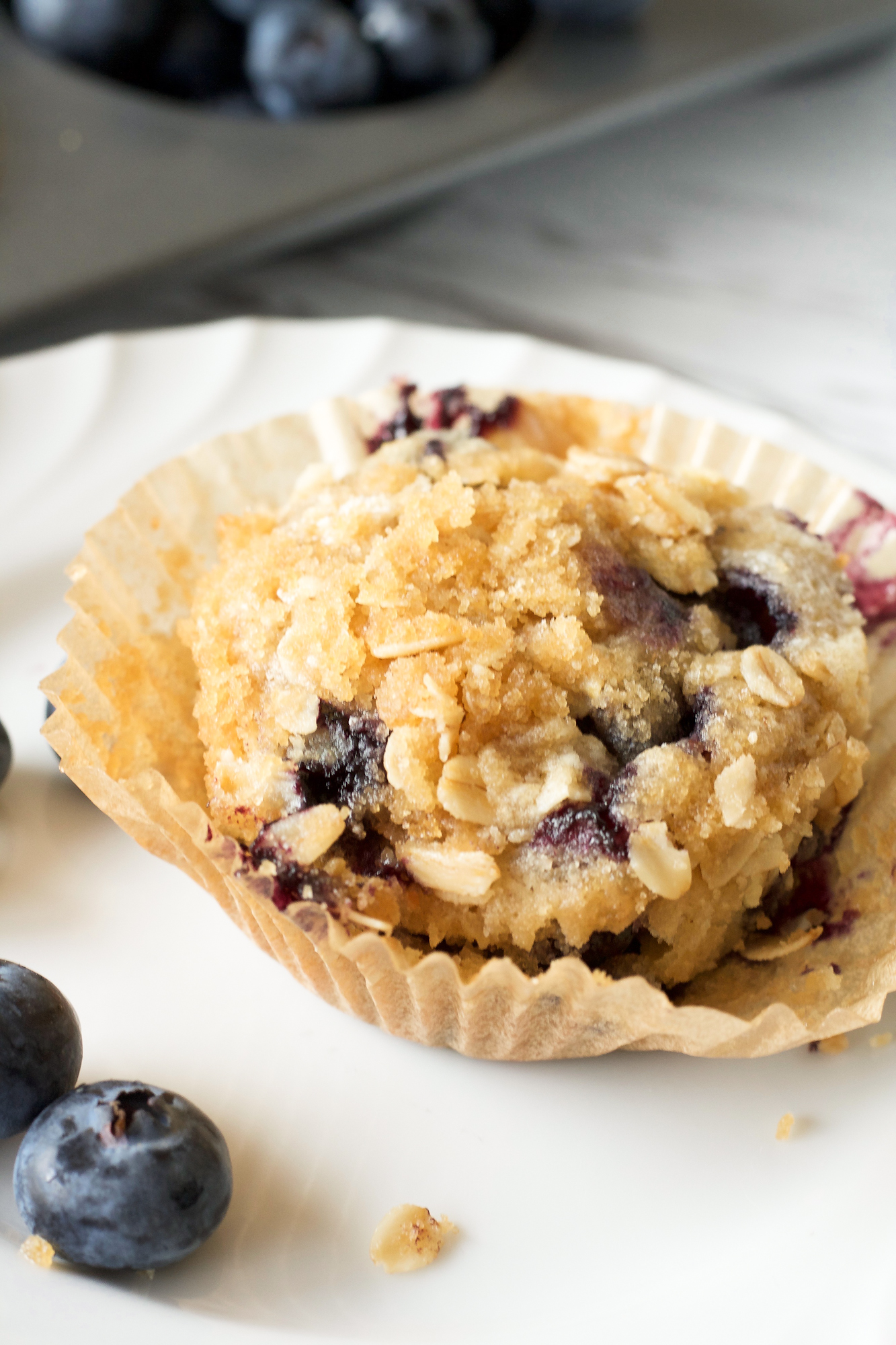 Sidebiew of a sourdough blueberry muffin topped with a oat streusel sitting on a white plate. The muffin paper is opening - Hostess At Heart
