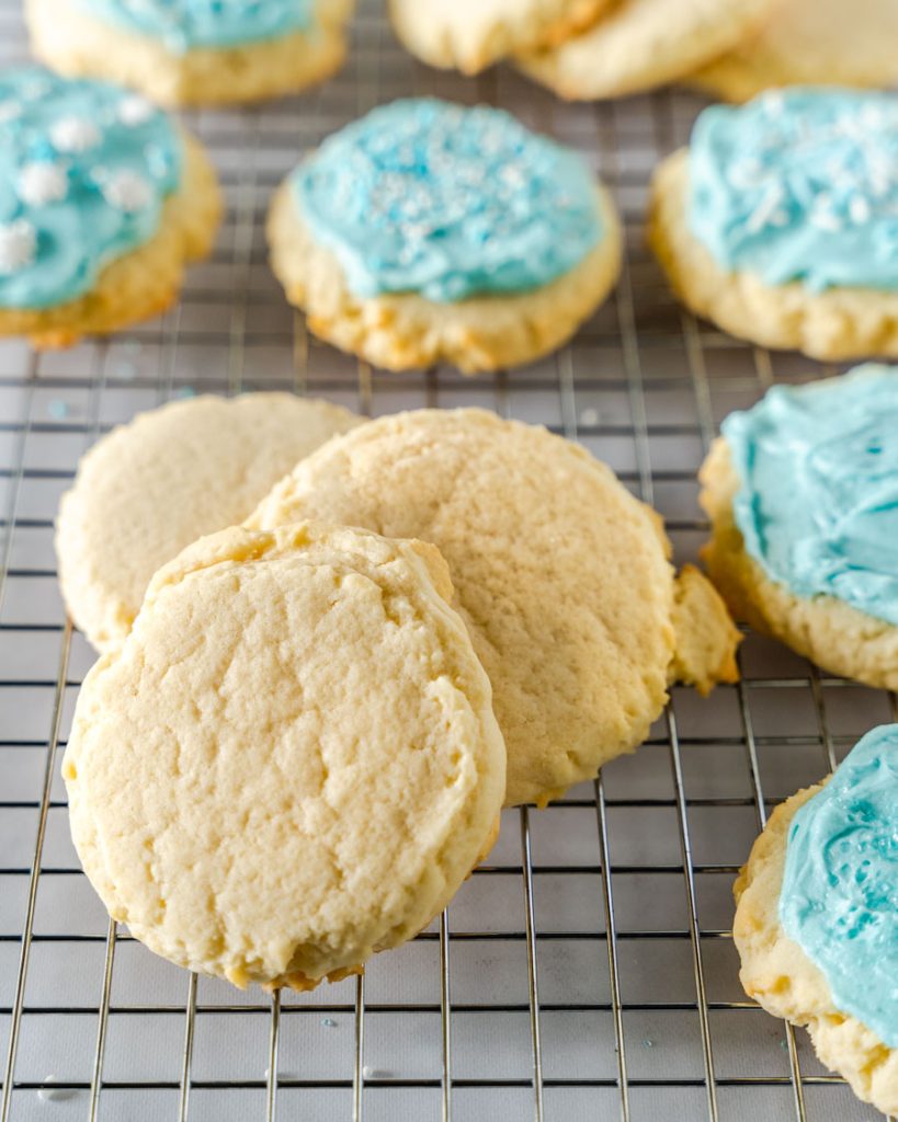 Three unfrosted sugar cookies sitting on a cooling rack in front of blue frosted cookies.
