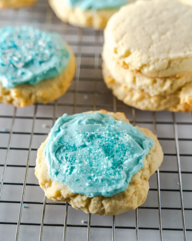 Angled view of a frosted sugar cookie and sprinkles sitting on a cooling rack in front of a stack of unfrosted cookies.