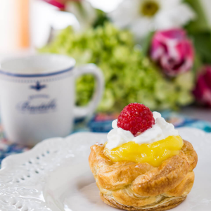 A baked puff pastry basket filled with lemon filling and topped with whipped cream and a raspberry.
