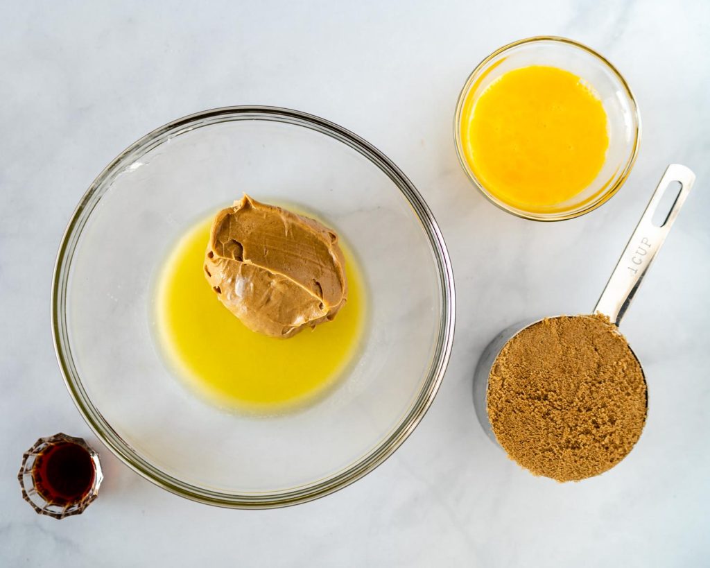 A bowl of melted butter with peanut butter added surrounded with a cup of brown sugar, vanilla, and egg.