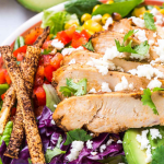 3/4 angle of a salad topped with sliced chicken, corn, red cabbage, Cotija, avocado and tortilla straws.