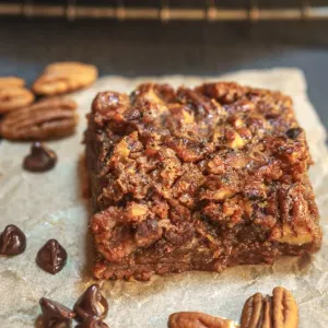 3/4 angle of a pecan pie brownie bar sitting on a piece of parchment paper surrounded by chocolate chips and pecans.