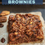 3/4 angle of a pecan pie brownie bar sitting on a piece of parchment paper surrounded by chocolate chips and pecans.