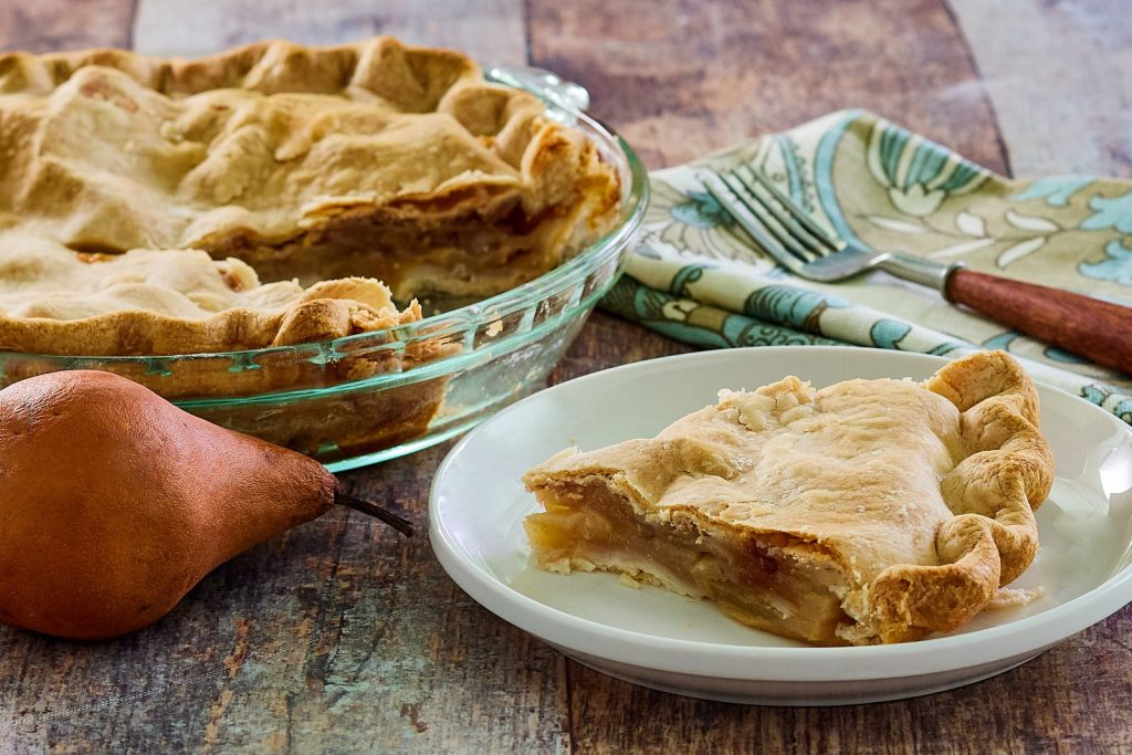 Easy pear pie recipe baked and on a plate. Single slice plated, full pie in background.