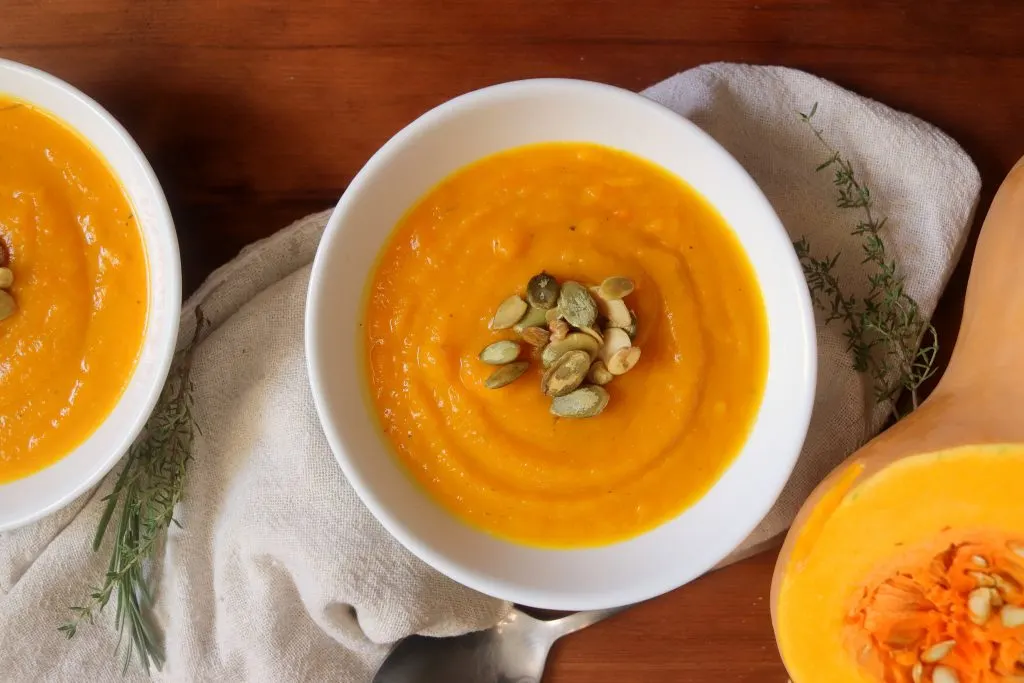 Top down view of creamy soup next to a butternut squash cut in half and herbs.