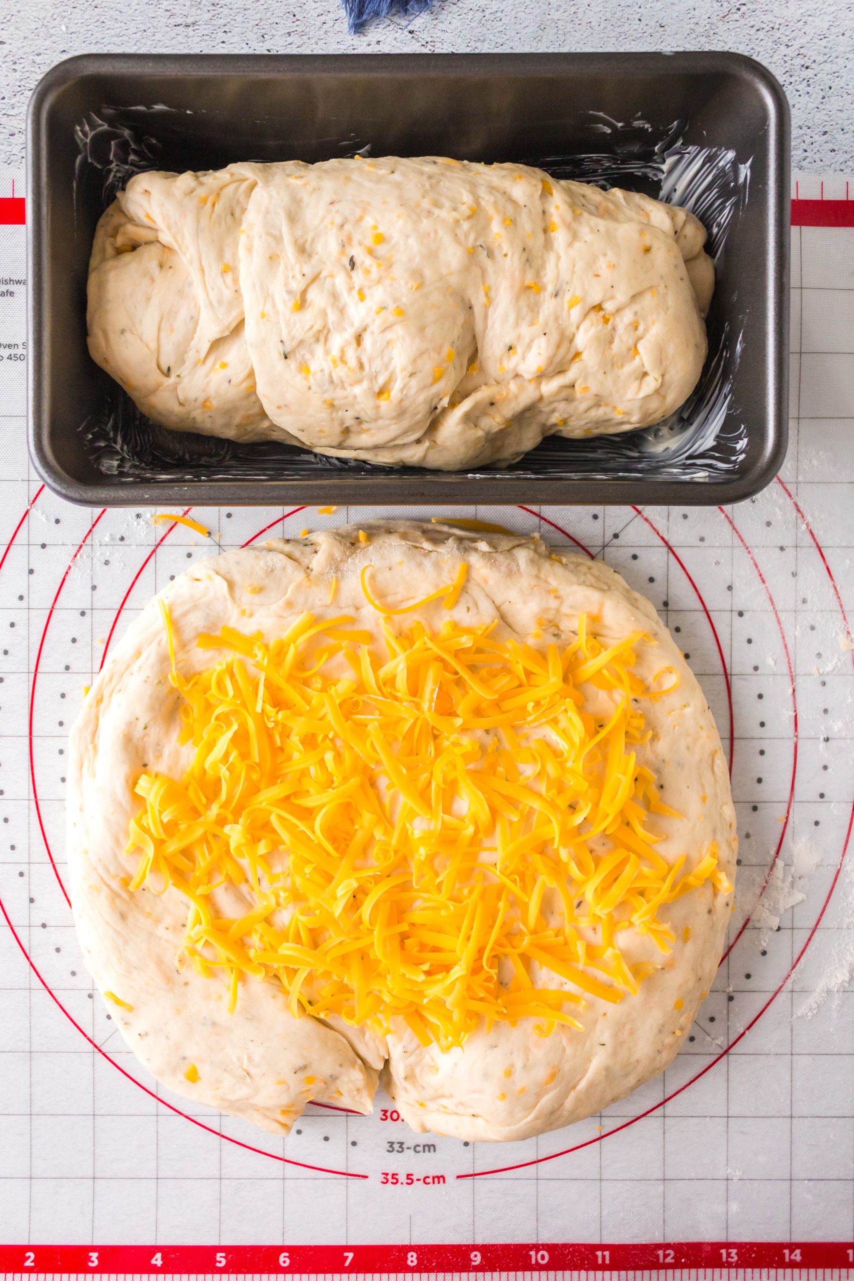 Top down view of a loaf pan filled with cheesy bread dough and another loaf rolled out on a pastry mat covered in cheese.