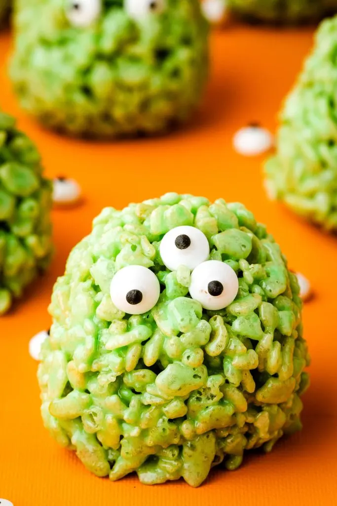 A green colored rice krispy treat shaped into a ball with candy eyeballs.
