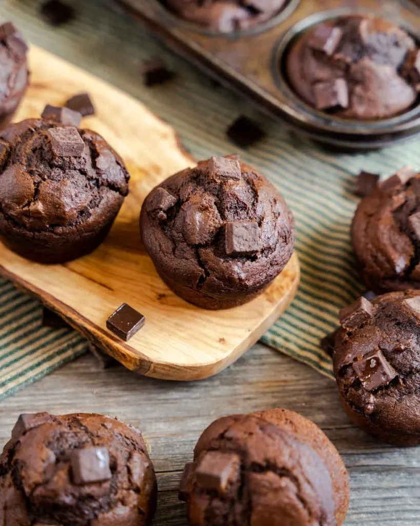 Two pumpkin muffins with chocolate sitting on a cutting board surrounded by more muffins.