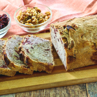 Side view of a sliced loaf of bread with bowls of dried cranberries and walnuts next to it.