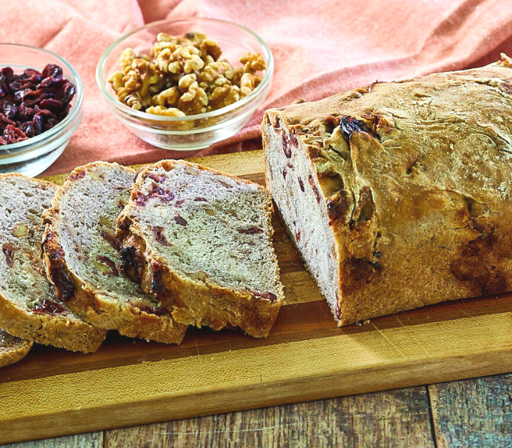 Side view of a sliced loaf of bread with bowls of dried cranberries and walnuts next to it.