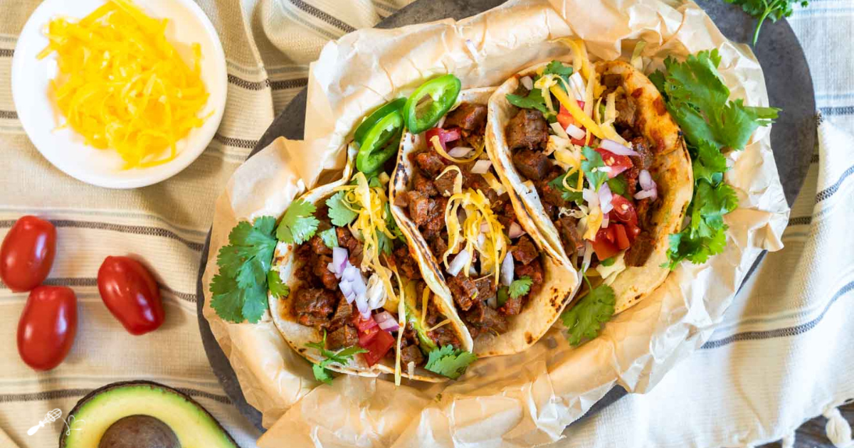 Top down view of beef brisket tacos in a basket surrounded with tomatoes, cheese, and avocado.