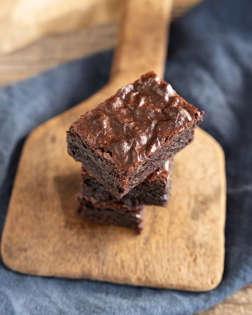 A dark chocolate brownie with a flaky top stacked on two more pieces of chocolate brownies.