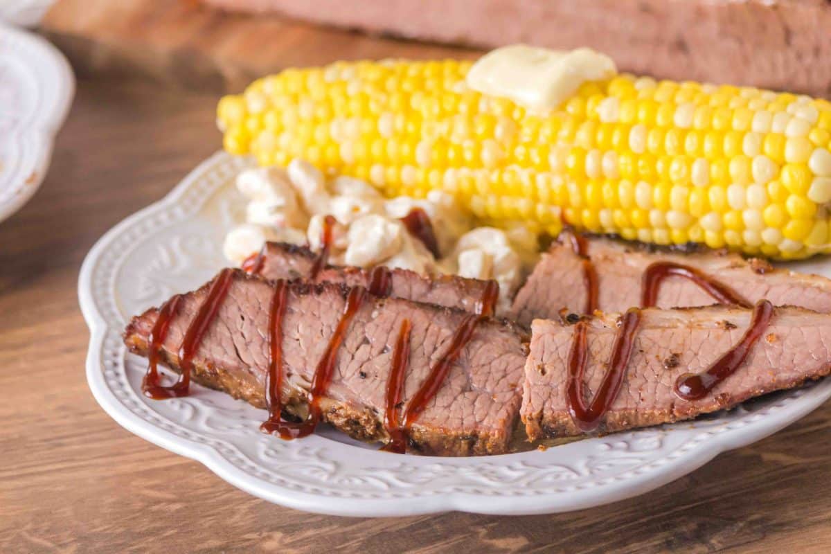 A plate filled with sliced beef brisket, potato Salad, and corn on the cob. Hostess At Heart
