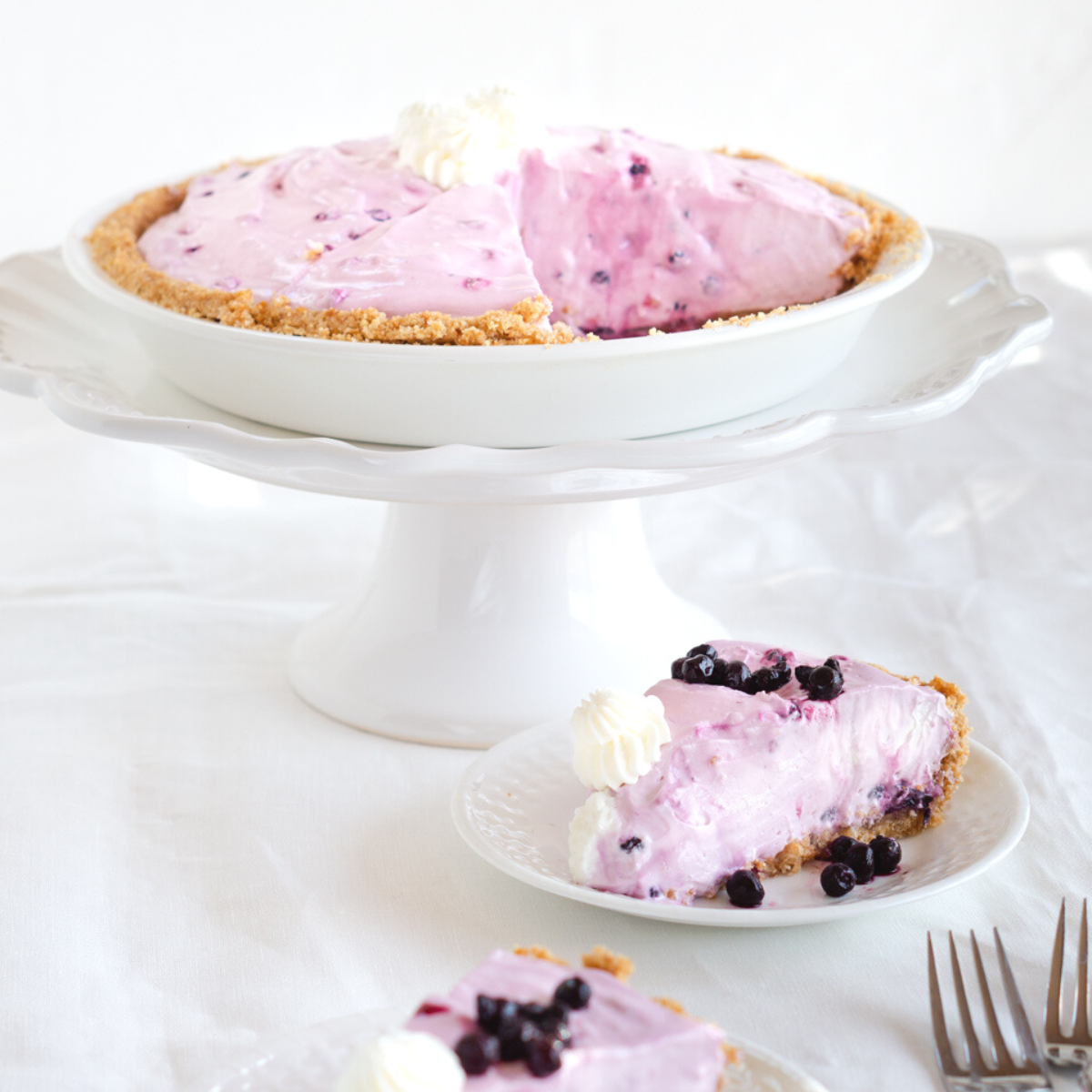 Two slices of blueberry ice box pie on plates surrounded by wild blue berries with a pie stand in the back holding the while pie.