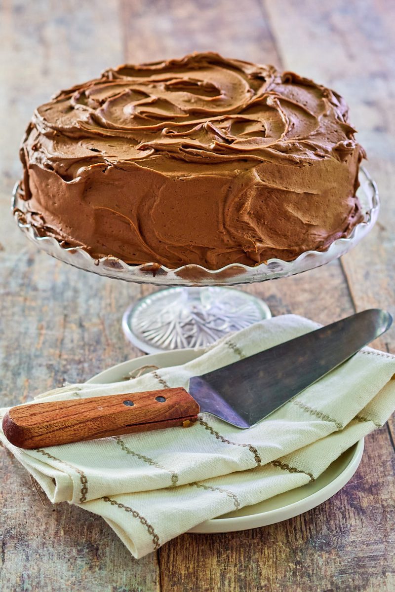 Side view of a chocolate mouse cake on a cake stand frosted with chocolate buttercream. A cake server sits in the front on a linen napkin placed over a serving plate.