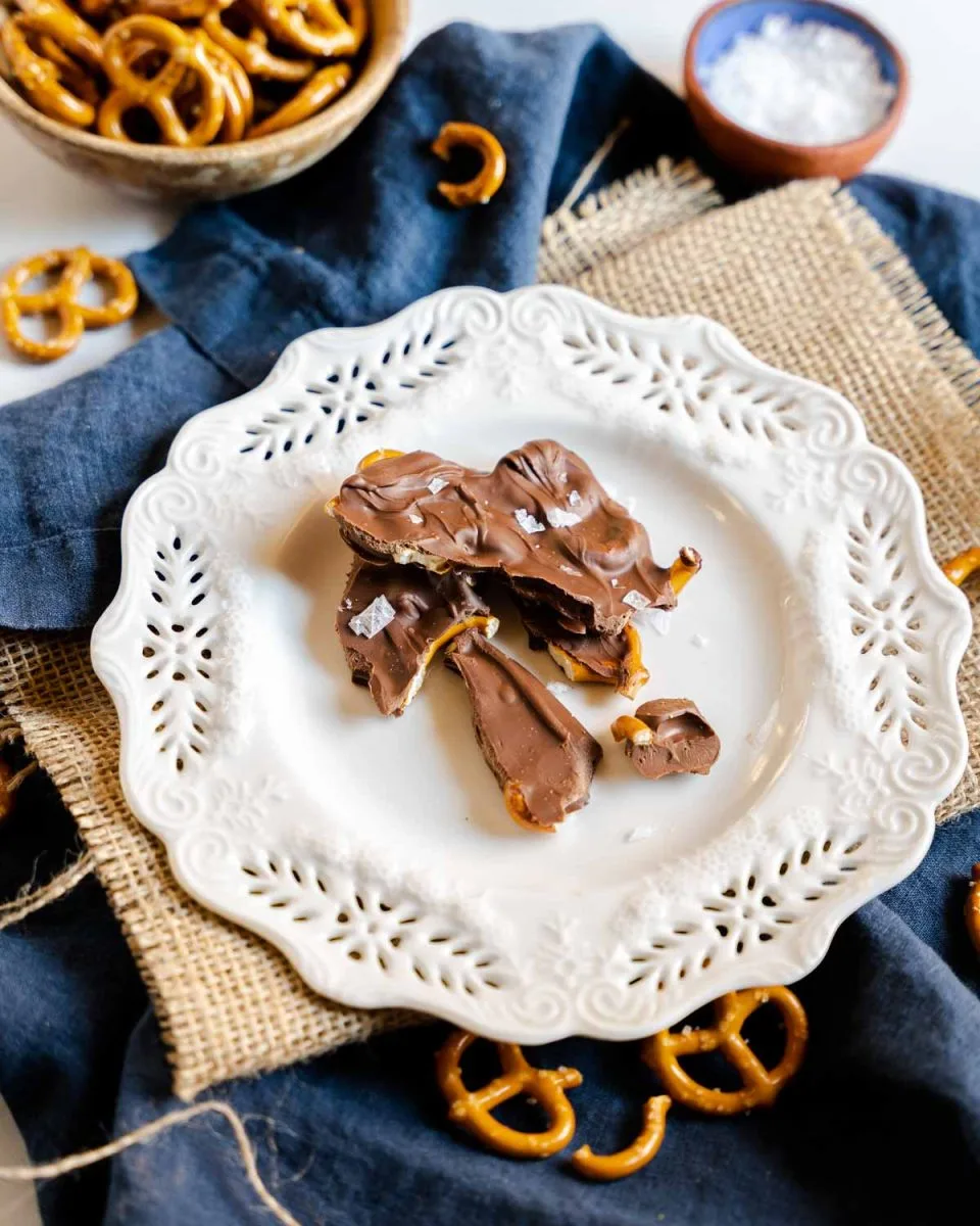 Angled view of broken chocolate bark covered pretzels sitting on a white plate. - hostess at heart