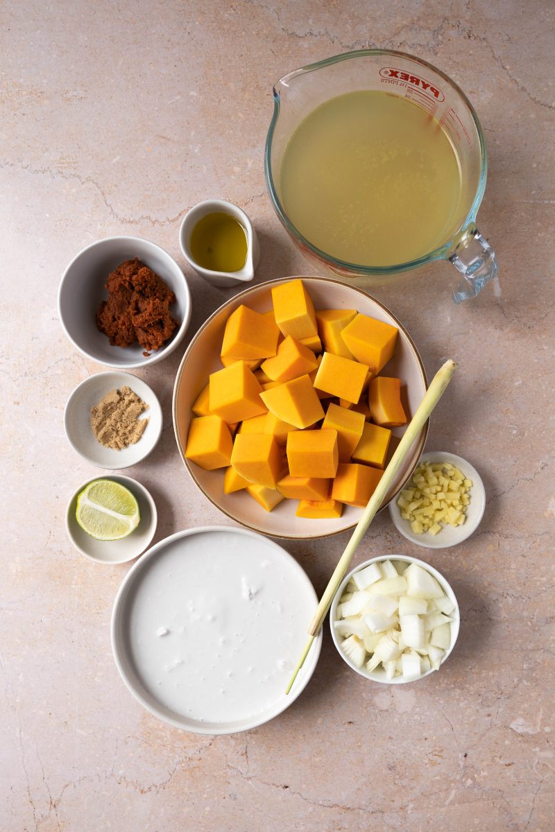 Ingredients used to make thai pumpkin soup with coconut milk including onion, pumpkin, lime, coconut milk, lemon grass, ginger, sugar, red curry paste, and oil.