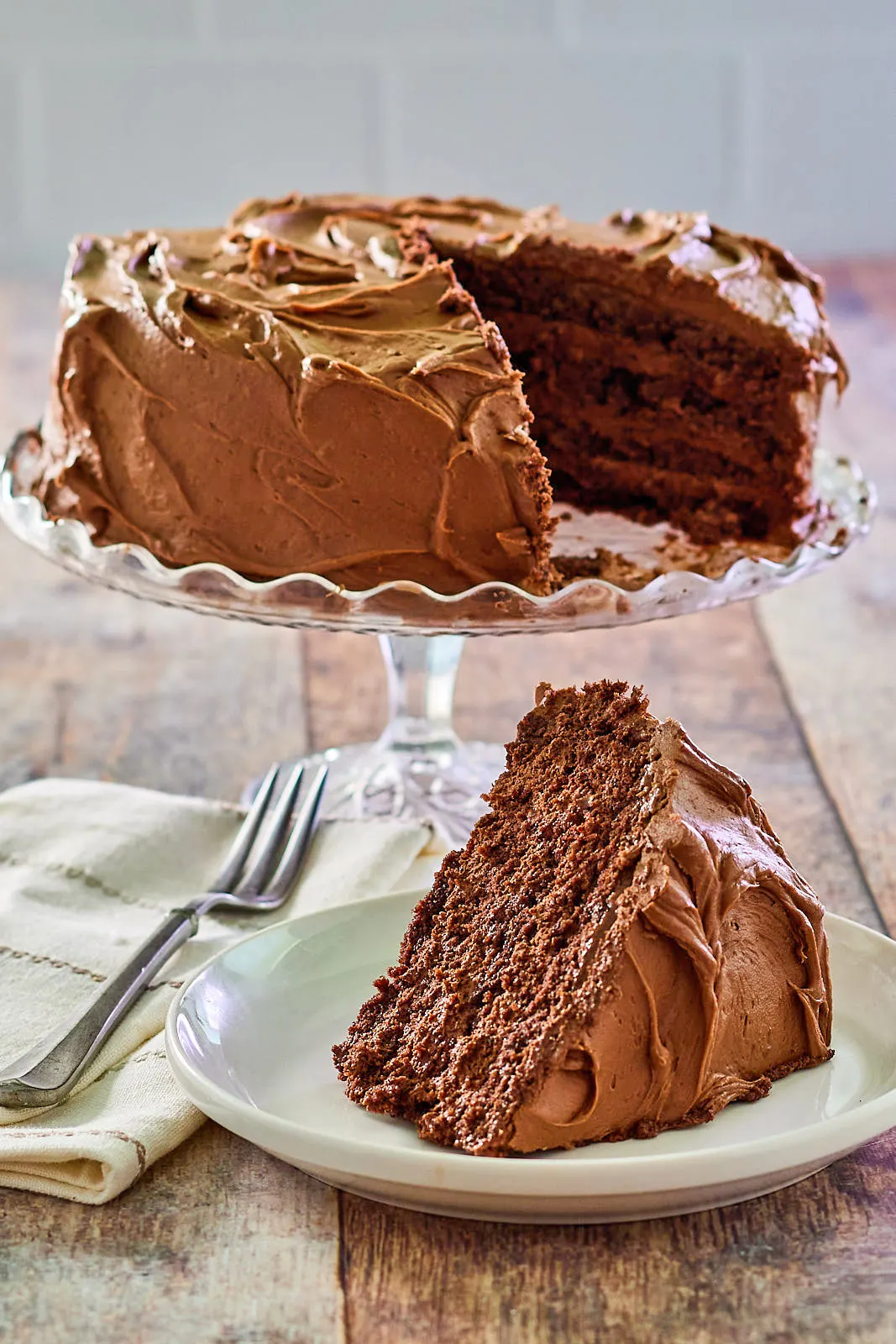 A tableview of a slice of chocolate cake with layers of chocolate mousse filling and frosted with chocolate buttercream. The sliced chocolate layer cake sits on a cake stand in the background. Hostess At Heart