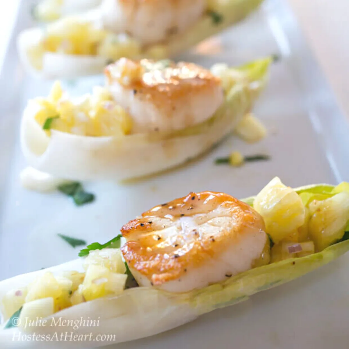 Sidebiew of a row of cooked scallops sitting on diced pineapple inside endive lettuce leaves on a white plate - Hostess At Heart