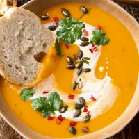 Top down view of pumpkin soup topped with cilantro, pepitas, diced red pepper and a whirl of cream. A slice of bread sits to the side.