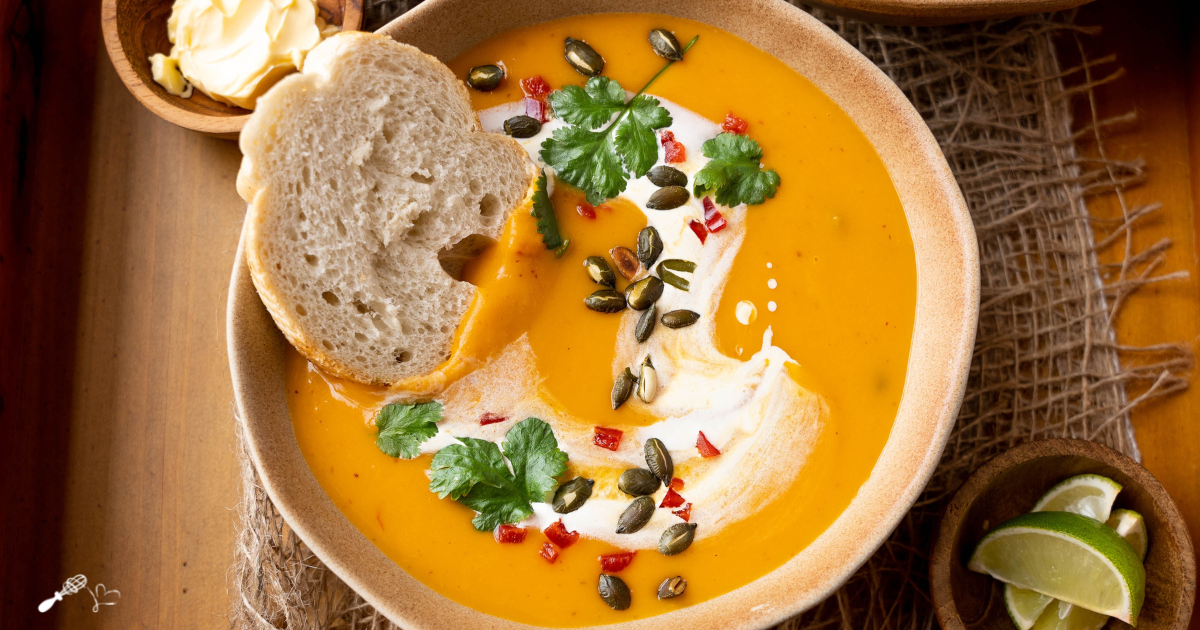 Top down view of pumpkin soup topped with cilantro, pepitas, diced red pepper and a whirl of cream. A slice of bread sits to the side.