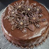 Angled view of a round layered cake covered in chocolate ganache on modeled chocolate flowers. Hostess At Heart