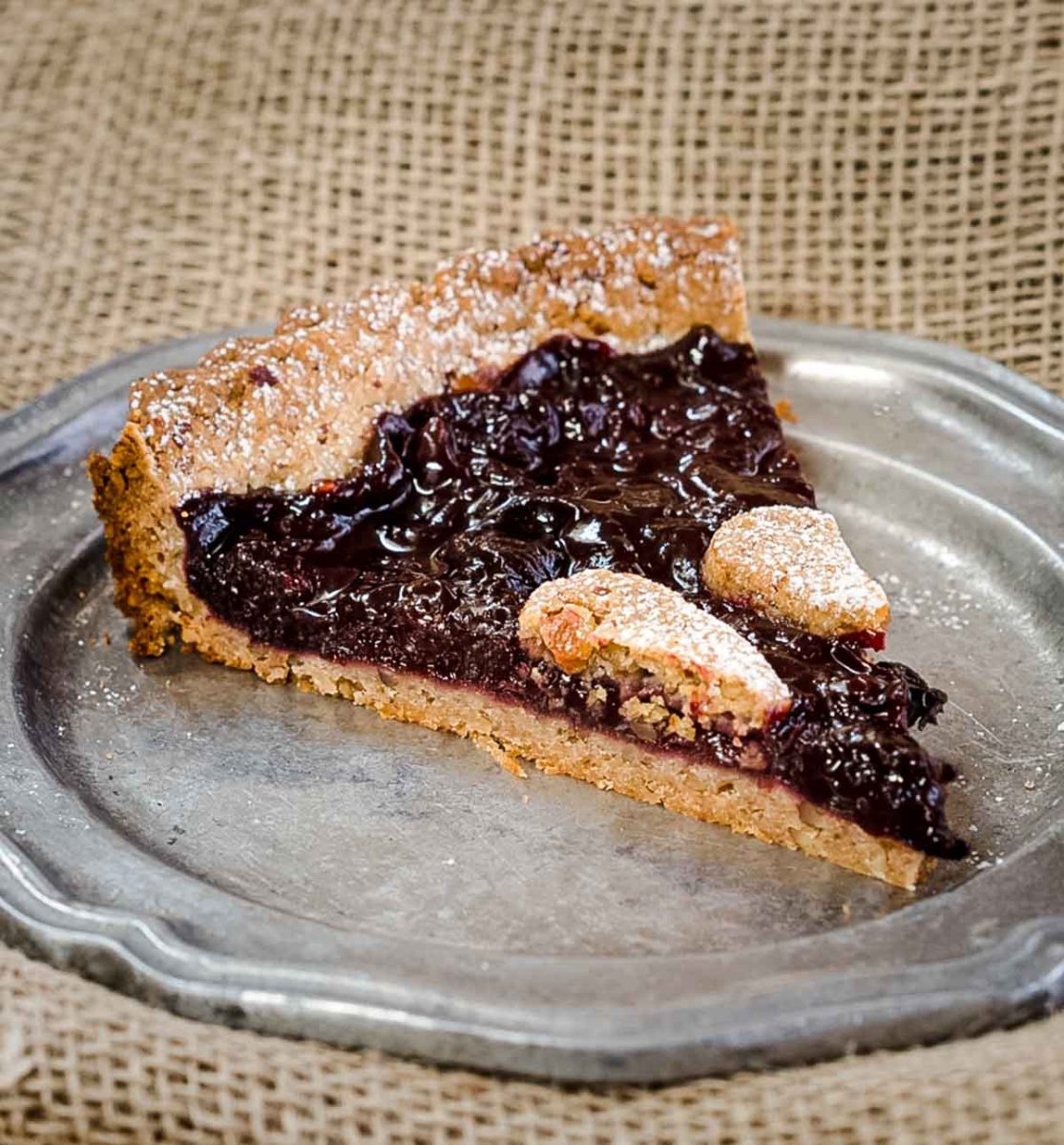 A slice of cherry torte dessert on a metal plate showing a thick dark cherry filling on a golden brown pecan crust. Hostess At Heart