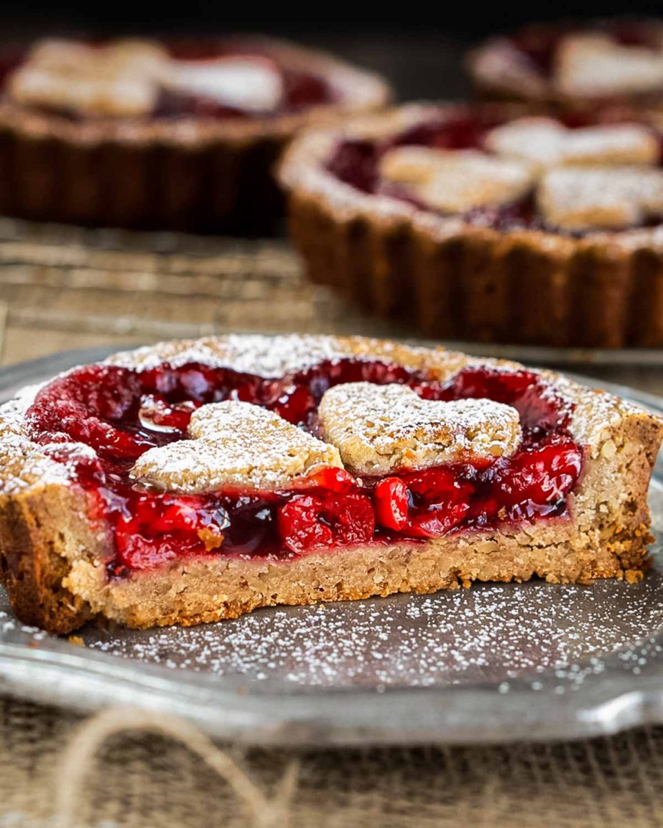 A front view of half of a cherry torte pie nestled in a pecan tart shell showing thick bright cherry filling in a browned crust and dusted with powdered sugar over the top. Hostess At Heart