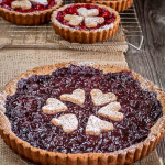 Angled view of a deep red cherry tart in a golden brown pecan crust with stamped crust hearts decorating the top. The whole thing is dusted with powdered sugar and sits in front of individual cherry tarts on a cooling rack. Hostess At Heart