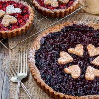 Angled view of a Sweet Cherry Tart sitting on a piece of decorative burlap in front of two individual cherry tarts sitting on a cooling rack. Hostess At Heart
