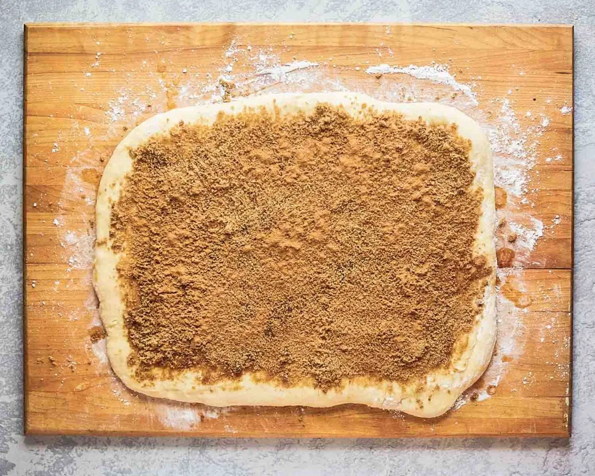 Homemade cinnamon roll dough rolled out on a cutting board and topped with a cinnamon and brown sugar filling. Hostess At Heart