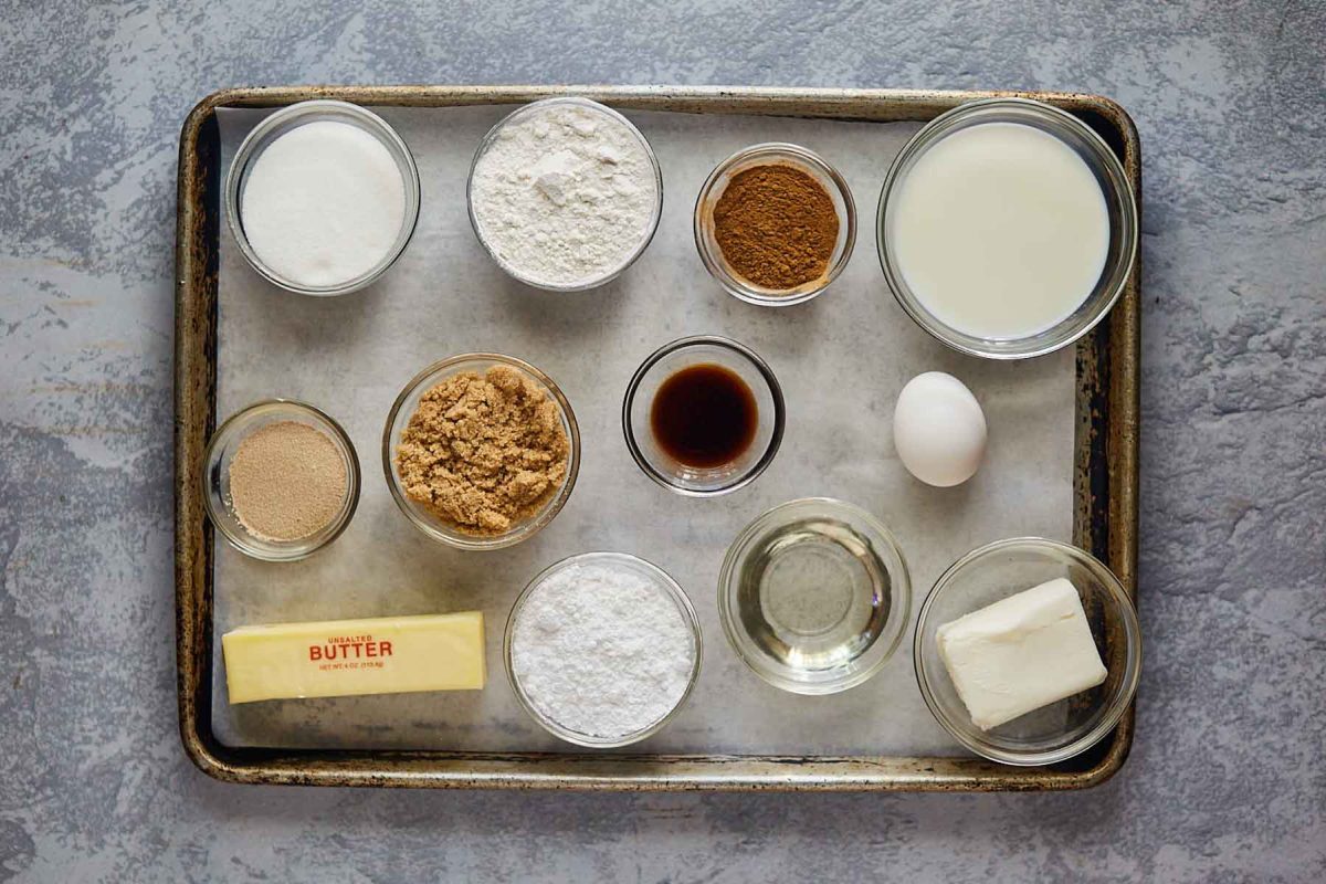Top down view of the ingredients used to make cinnamon rolls from scratch including shortening, sugar, milk, butter, vanilla, egg, flour, and yeast