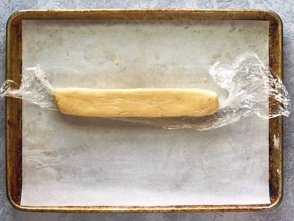 A log of cookie dough wrapped in plastic wrap on a parchment paper-lined cookie sheet. - Hostess At Heart