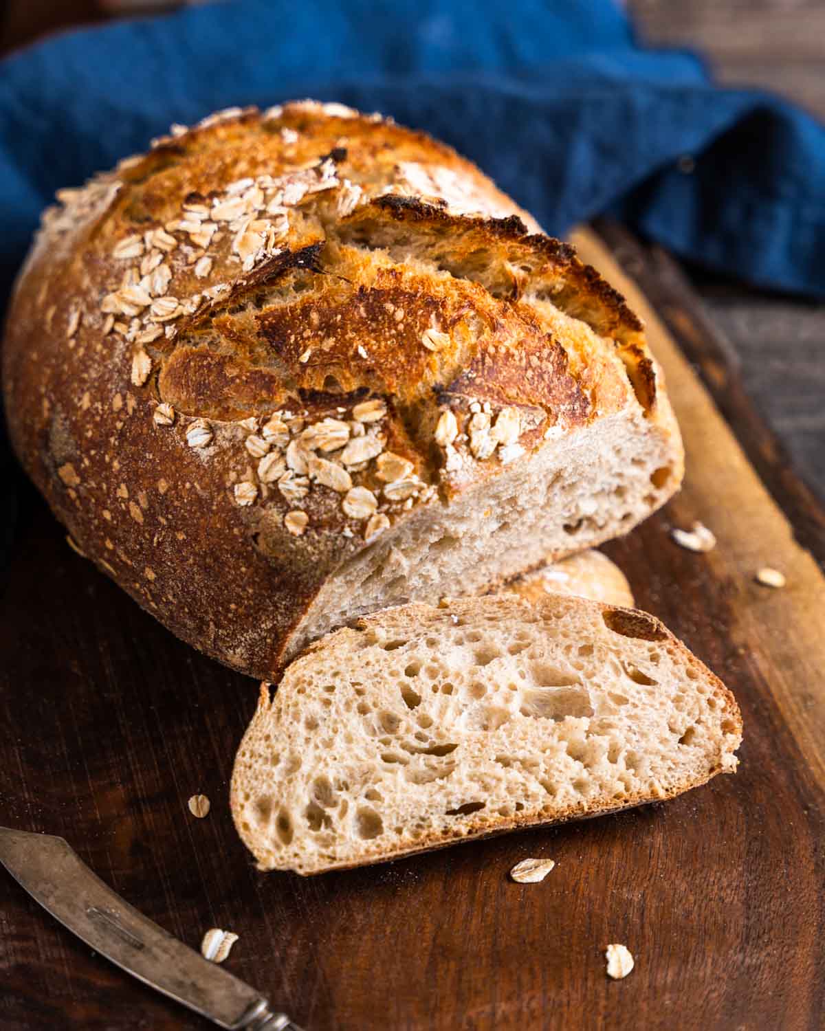 A golden brown loaf of rye sourdough bread topped with oats on a cutting board - hostess at heart