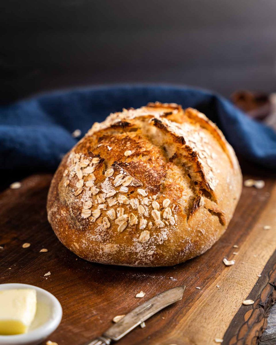 A golden brown loaf of rye sourdough bread topped with oats on a cutting board - hostess at heart