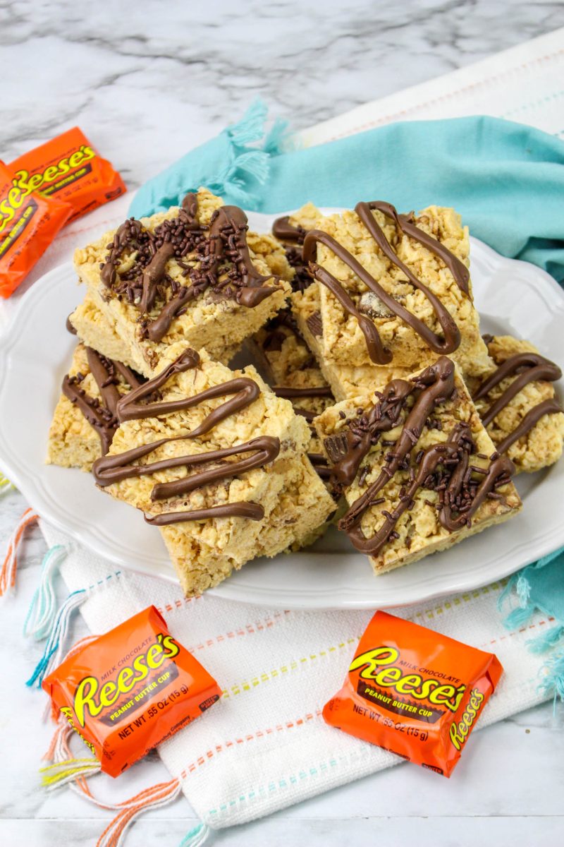 Peanut Butter Rice Krispies Treats on a plate from a more distant view and Reese's peanut butter cups in the background.