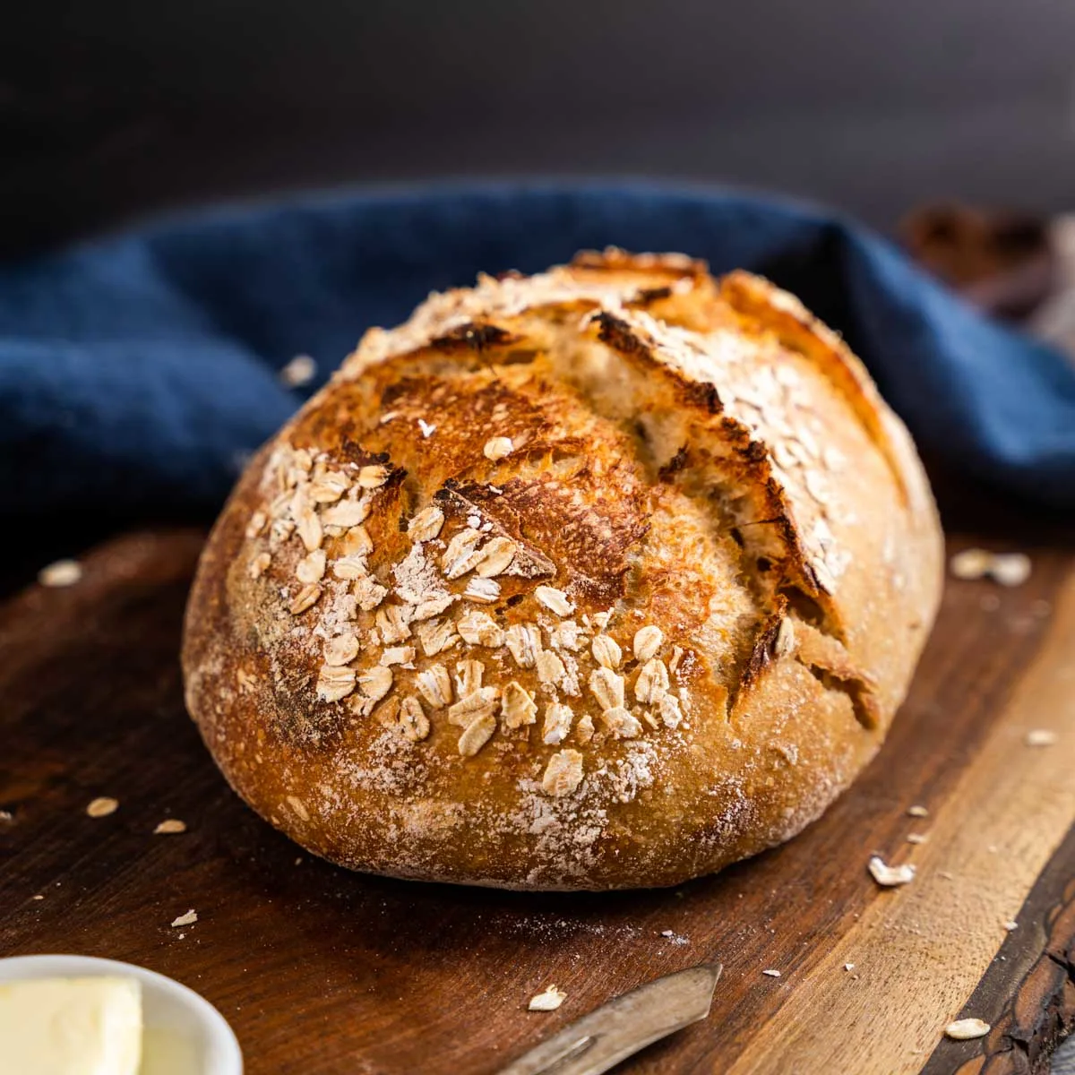 A golden brown loaf of rye sourdough bread topped with oats on a cutting board - hostess at heart