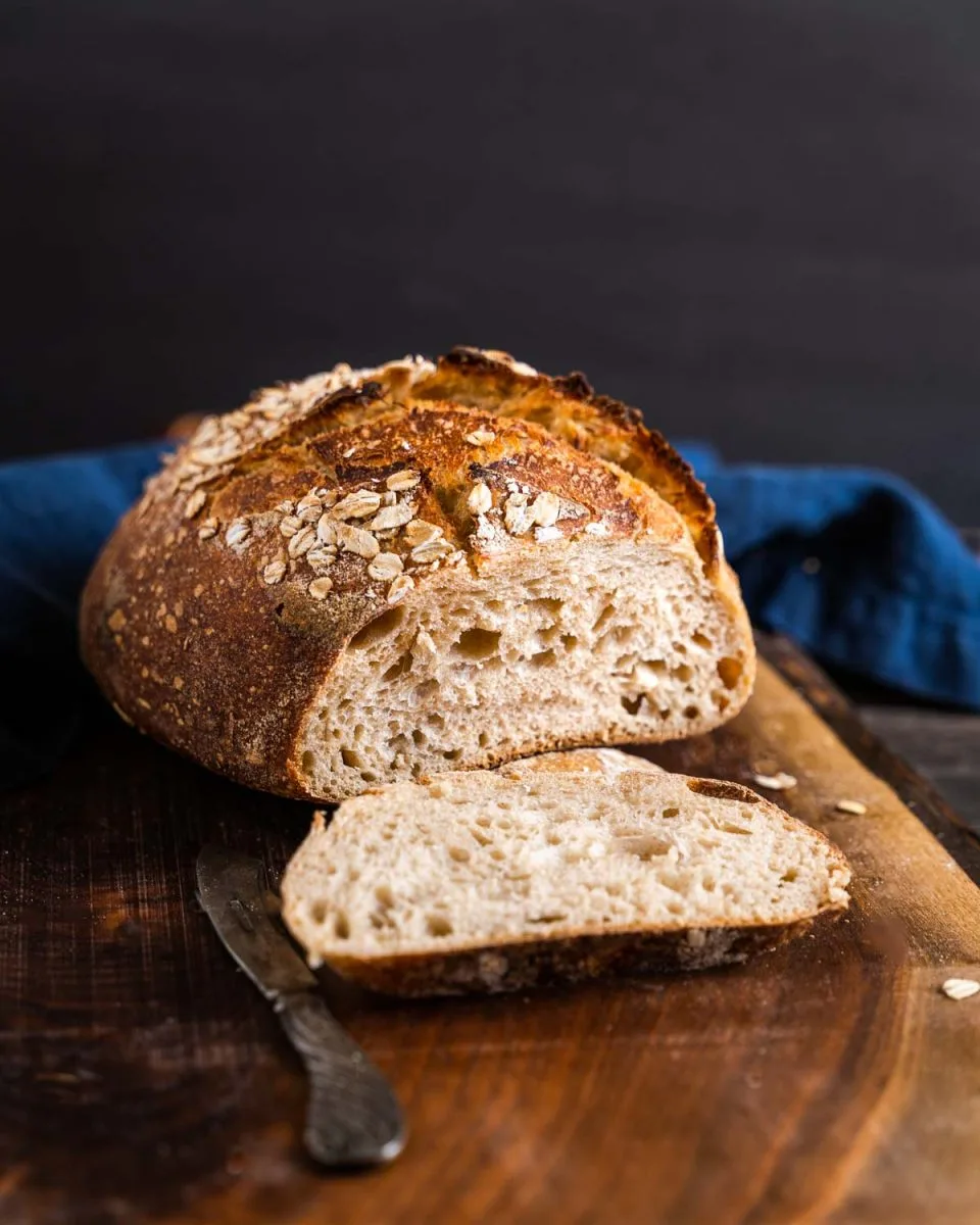 Front view of a sliced loaf of light rye bread topped with oats. A slice lies flat in front of the loaf - hostess at heart