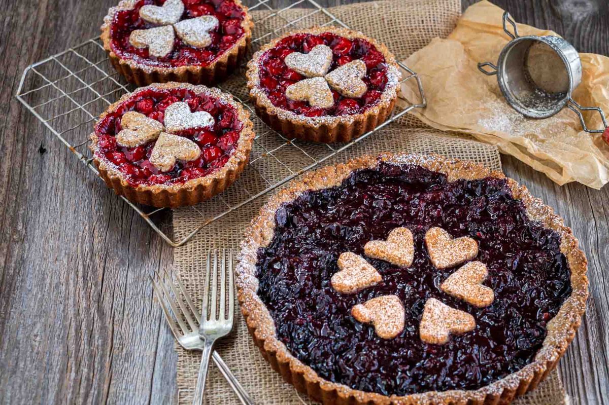 Angled view of a large Sweet Black Cherry Tart sitting in front of individual Cherry Pie Filling tarts on a cooling rack. Hostess At Heart