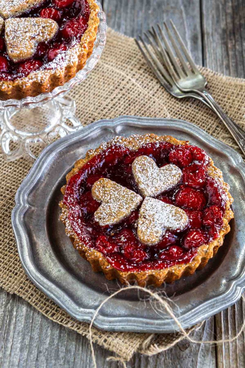Angled view of a cherry tart topped with pastry hearts sits on a metal plate over a piece of burlap. Hostess At Heart