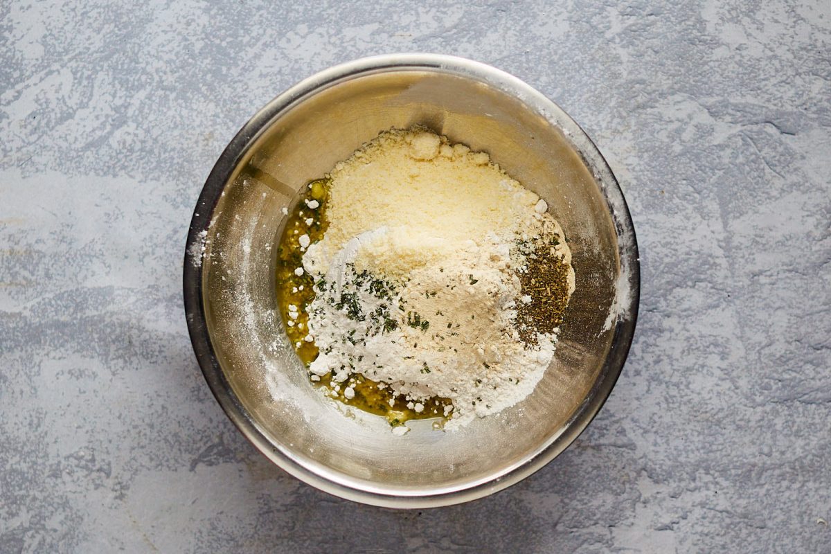 Top down view of dry bread ingredients used in an easy focaccia bread recipe including flour, cheese, and herbs. Hostess At Heart