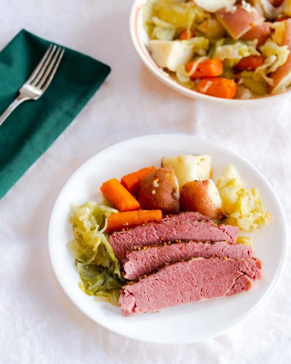 Sliced Instant Pot Corned beef sitting on a white plate surrounded by cooked cabbage, carrots, and Potatoes - Hostess At Heart