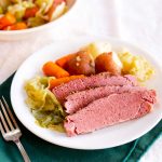 Angled view of sliced corn beef on a platter surrounded by cooked carrots, cabbage and potatoes - Hostess At Heart