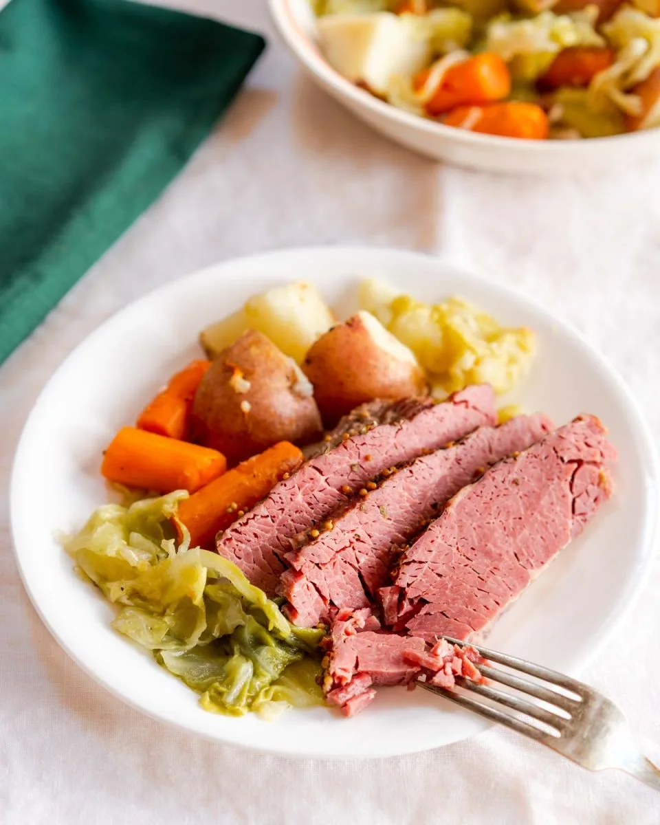 Angled view of a fork flaking a slice of corned beef on a plate surrounded by veggies - Hostess At Heart