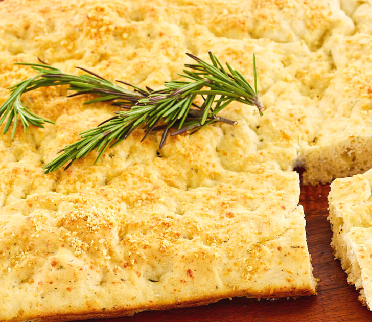 Angled view of focaccia bread topped with rosemary and parmesan cheese sitting on a cutting board - Hostess At Heart