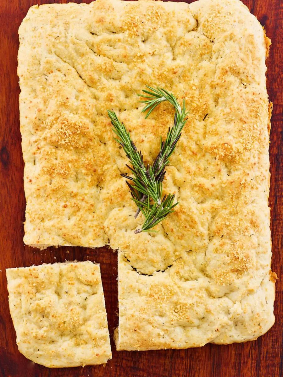 Top down view of baked rosemary foccacia bread sitting on a cutting board with a piece sliced off. Hostess At Heart