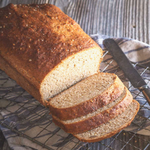 Angled view of a loaf of Honey multigrain bread sitting on a cooling rack with the front cut into 3 slices - Hostess At Heart